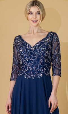 A woman smiles against a beige background, wearing the Amelia Couture 7046 sleeved long formal dress in navy blue, featuring intricate beadwork and sheer three-quarter sleeves.