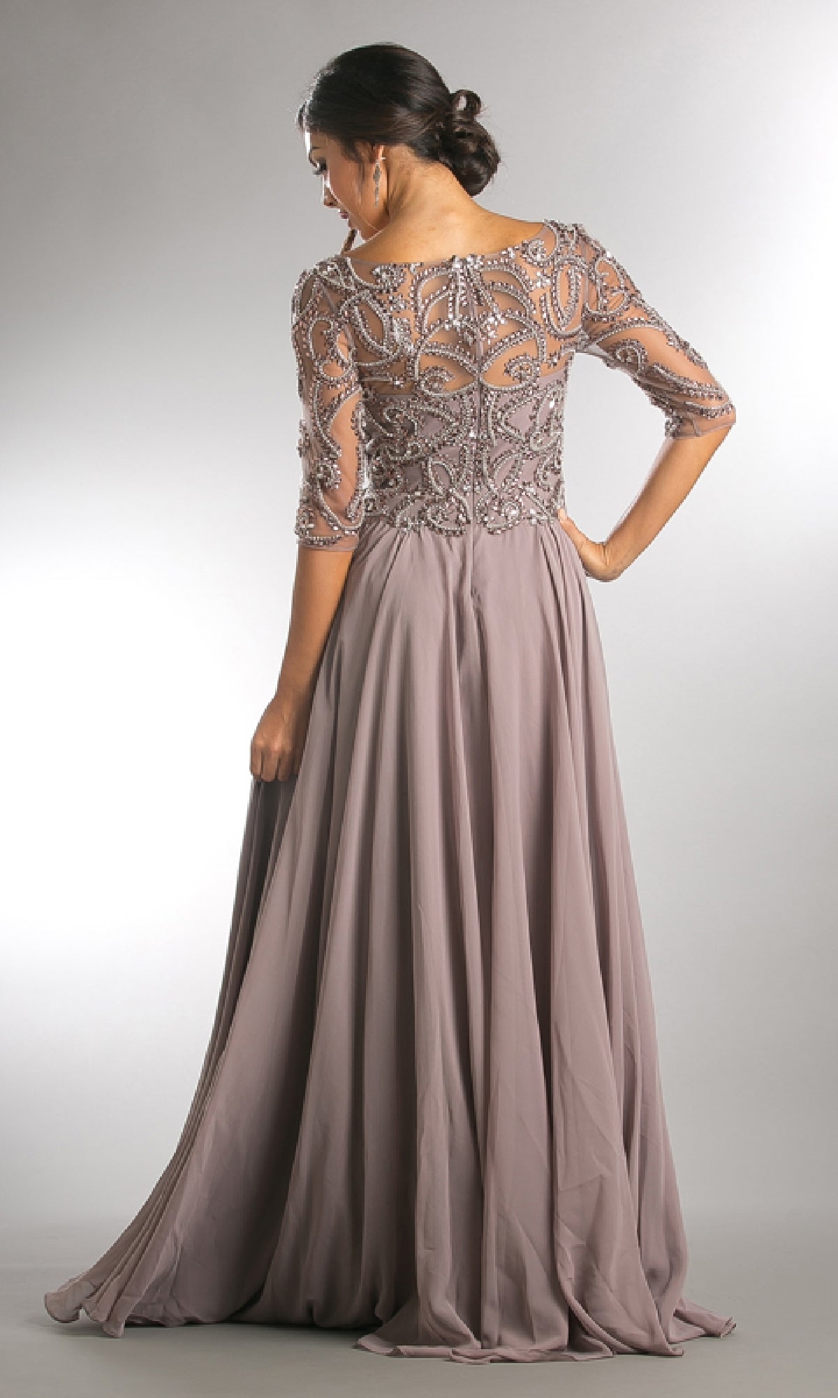 A woman stands with her back to the camera in the Amelia Couture 7046 sleeved long formal dress, featuring a floor-length mauve design with a beaded, semi-sheer bodice and flowing skirt.