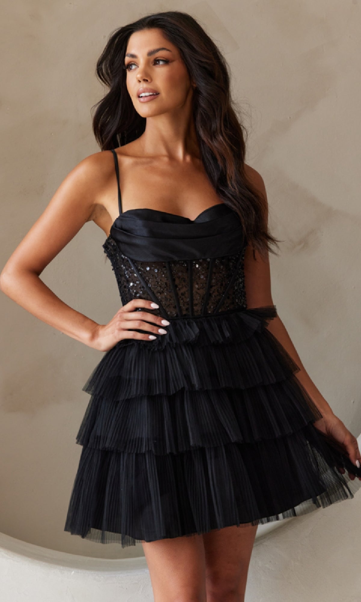 A woman stands against a plain light wall wearing the Beaded-Bodice Short Ruffled Hoco Dress BZ9023S, a black strapless mini with a fitted bodice and tiered ruffled skirt.