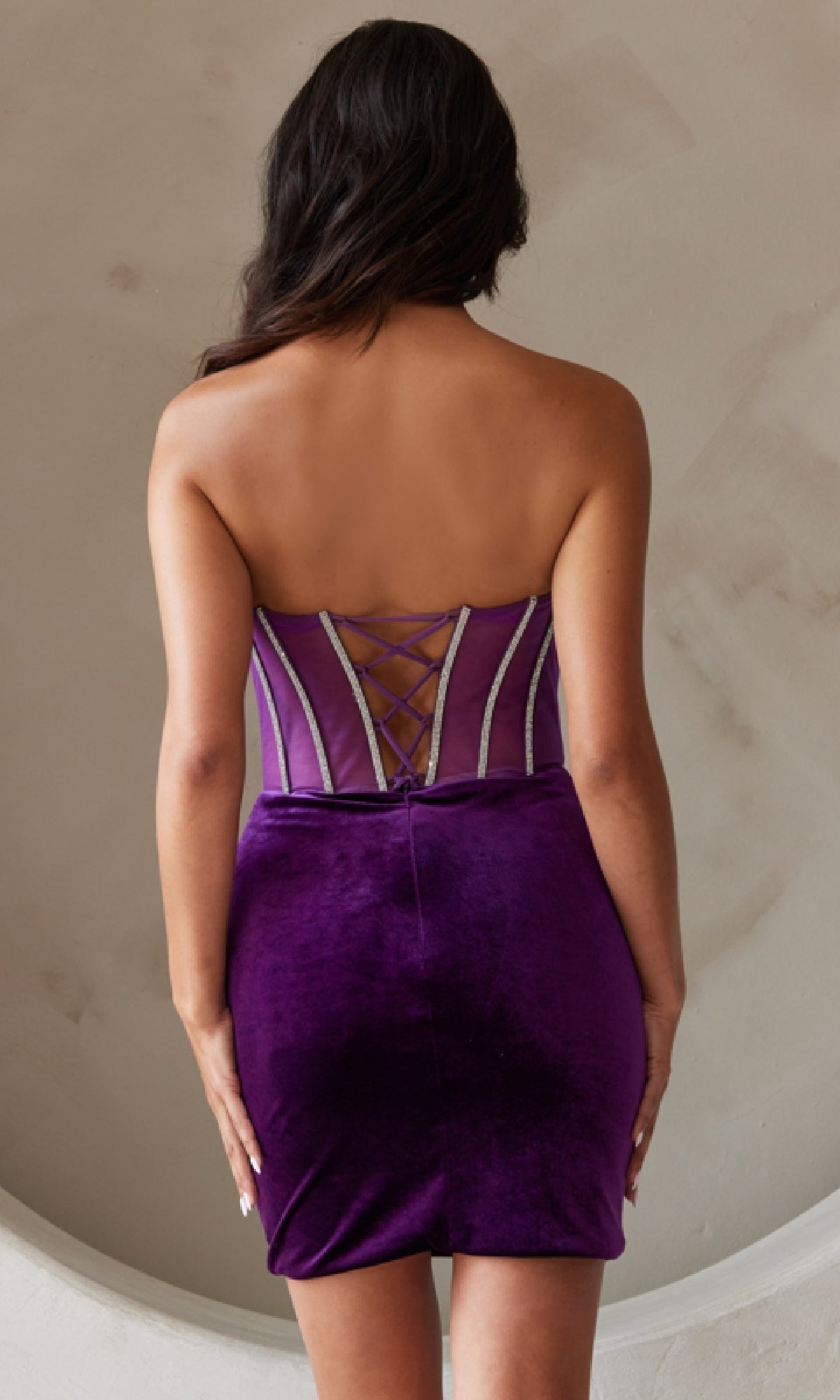A woman stands with her back to the camera, wearing the Strapless Short Velvet Homecoming Dress 5056S featuring a beaded corset bodice and lace-up corset back.