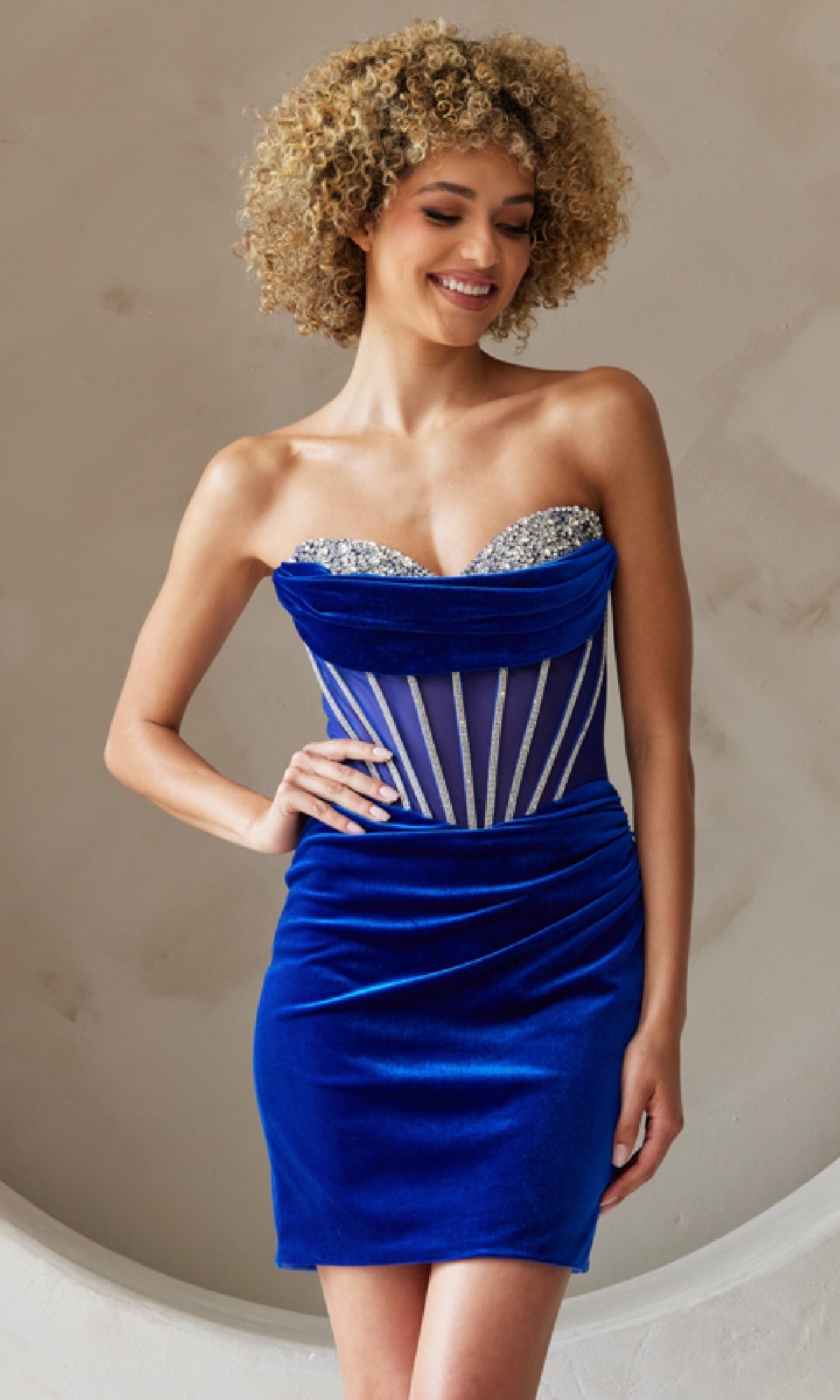 A woman in the Strapless Short Velvet Homecoming Dress 5056S, featuring a beaded corset bodice, stands against a light wall, smiling with one hand on her hip.