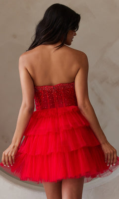A woman with long dark hair wears the Strapless Short Ruffled A-Line Hoco Dress BZ9999S, featuring a beaded bodice and layered red tulle, shown from the back against a neutral wall.