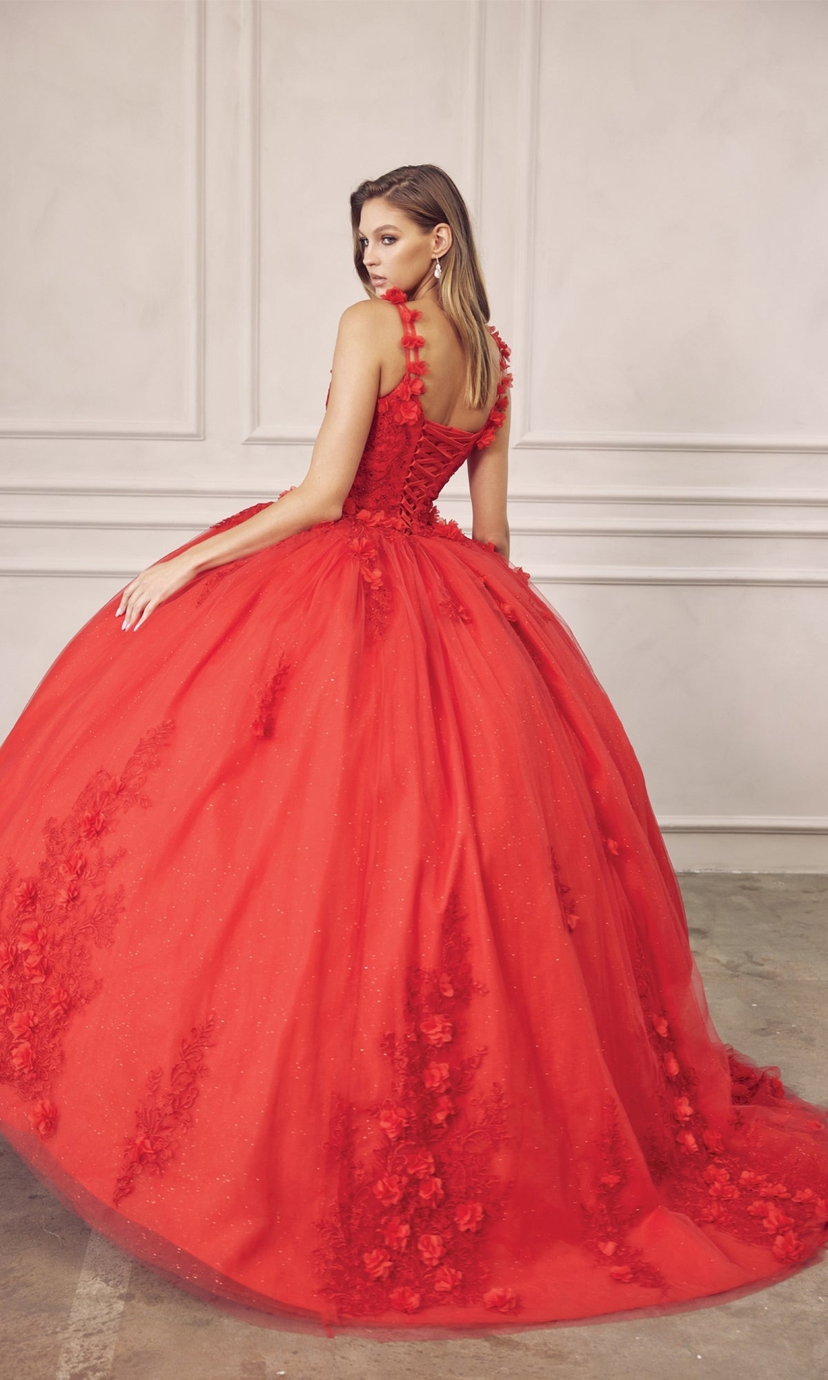 A woman wearing the Juliet 1437R Red Ball Gown Quinceañera Dress, featuring beaded embroidery and a voluminous skirt, stands sideways in a minimalist room with white panel walls.