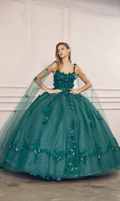 A woman wears the Juliet 1436 Glitter Quinceañera Dress with cape, featuring beaded embroidery, floral appliqués, a voluminous tulle skirt, and a matching sheer cape, set against a white-paneled wall.