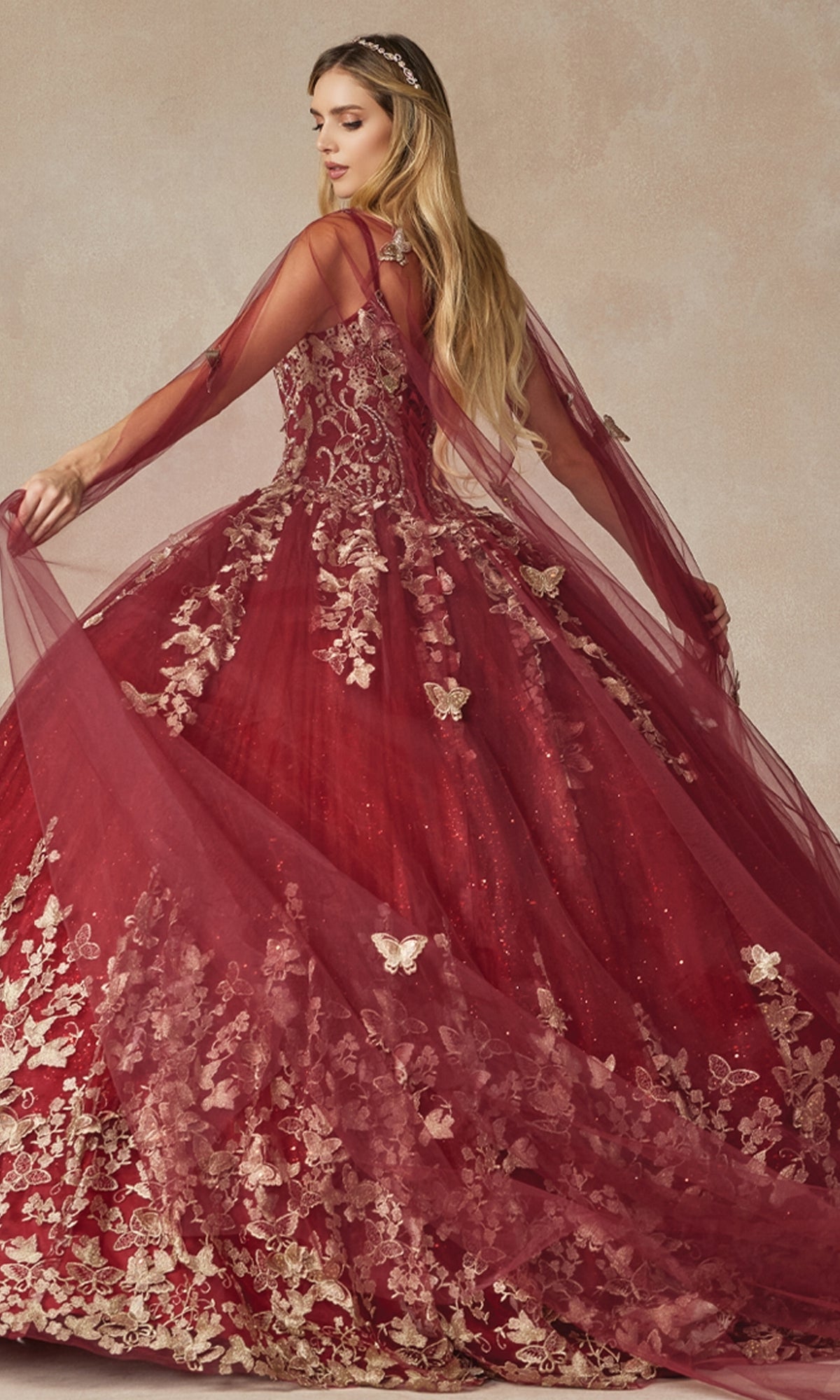A woman poses in the Juliet 1442 Quinceañera Dress, a red ball gown featuring floral and butterfly appliqués, with its sheer cape elegantly flowing behind her.