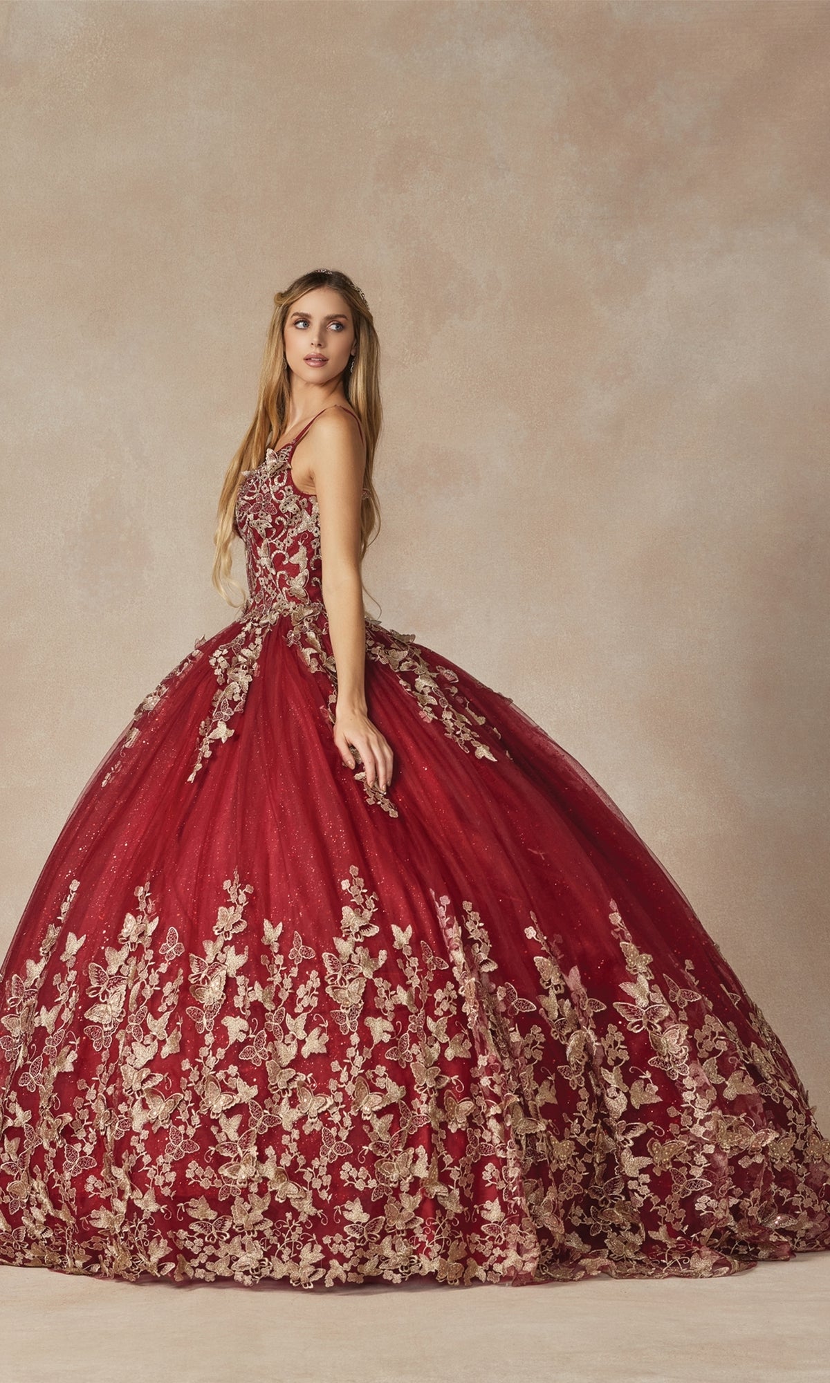 A woman stands against a neutral background wearing the Juliet 1442 Quinceañera Dress with a sheer cape, featuring sleeveless burgundy design and gold floral embroidery.