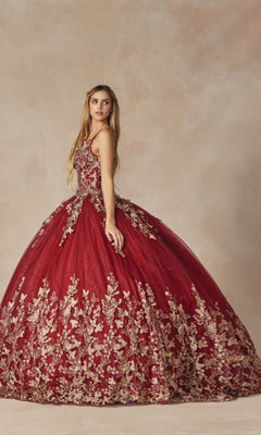 A woman stands against a neutral background wearing the Juliet 1442 Quinceañera Dress with a sheer cape, featuring sleeveless burgundy design and gold floral embroidery.