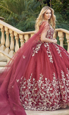 A woman stands on stone steps outdoors, surrounded by greenery, wearing the Juliet 1442 Quinceañera Dress with gold floral embroidery, deep red hue, and a long sheer cape.