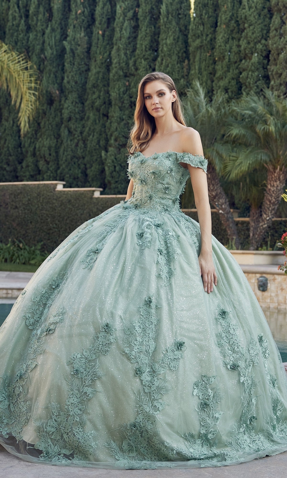 A woman stands outdoors in the Juliet 1445 Sage Green Quinceañera Dress Ball Gown, featuring an off-the-shoulder design, floral appliqués, and a full tulle skirt. Tall trees and lush greenery create a vibrant backdrop.