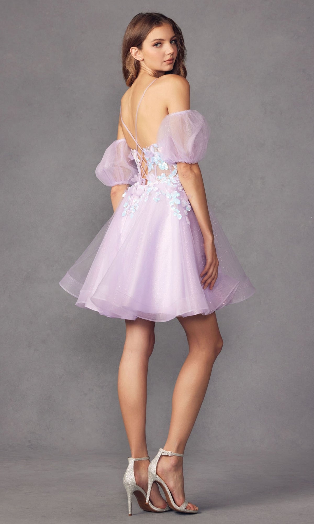 A woman wears the Puff-Sleeve Short Butterfly Homecoming Dress 901, a light purple babydoll dress with puff sleeves, floral appliqués, and an open lace-up back, paired with silver heels against a gray background.