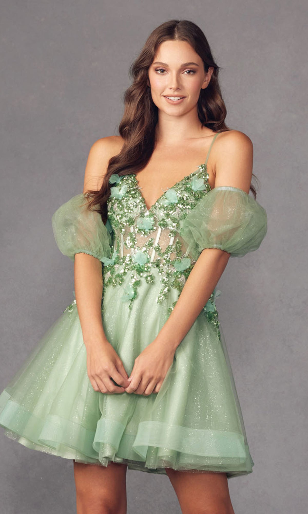 Woman wearing the Puff-Sleeve Short Butterfly Homecoming Dress 901, a light green babydoll style with sheer puff sleeves, floral appliqué, butterfly details, and glitter accents, standing against a plain gray background.