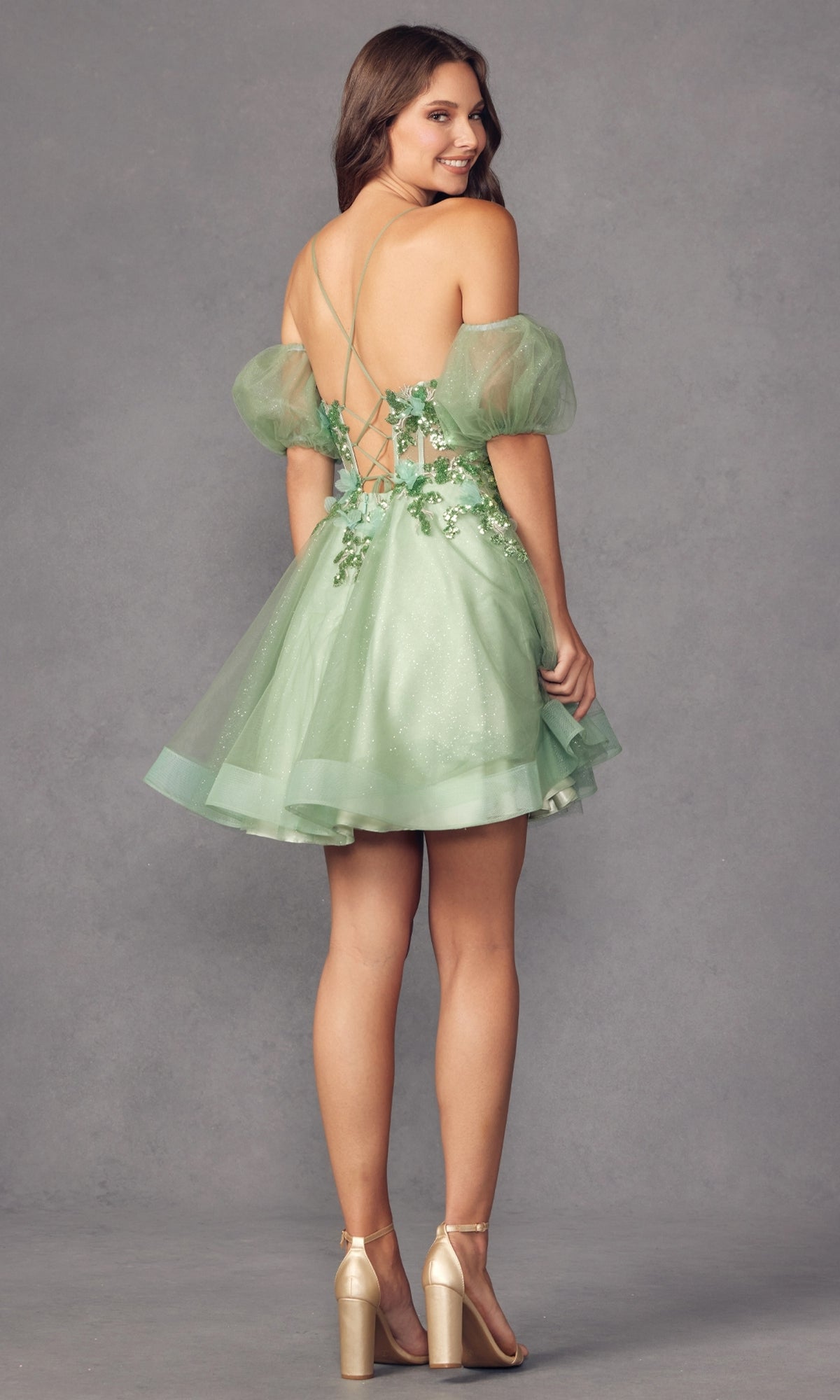 A woman models the Puff-Sleeve Short Butterfly Homecoming Dress 901, featuring light green fabric, puffy sleeves, butterfly appliqué, sheer waist, and an open lace-up back—perfect for making a statement against the gray background.