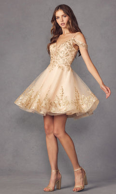 A woman stands against a gray background wearing the Glitter-Print Short Babydoll Homecoming Dress 903 with off-shoulder champagne layers, gold embroidery, sheer accents, and clear-strap block-heeled sandals.