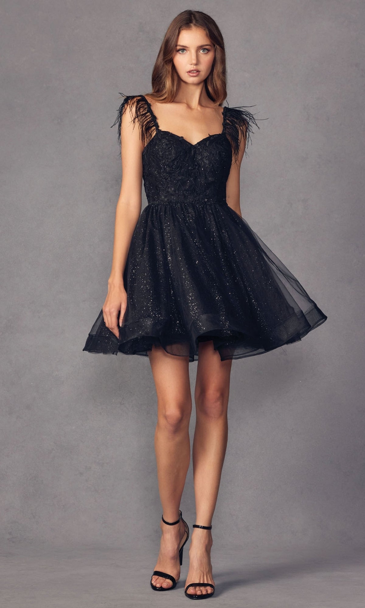 A woman models the Short Babydoll Homecoming Dress with Feathers 881, featuring a glitter finish, feathered shoulder accents, and black high-heeled sandals, set against a plain gray background.