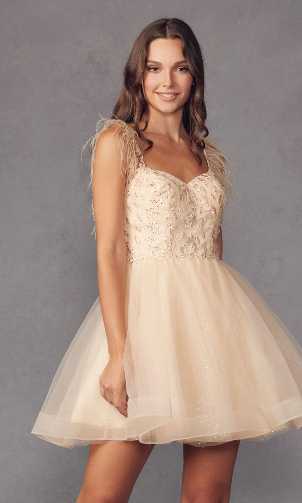 A woman wears the Short Babydoll Homecoming Dress with Feathers 881—a knee-length, light beige tulle dress with feathered shoulder straps and an embroidered sweetheart bodice—against a plain gray background.