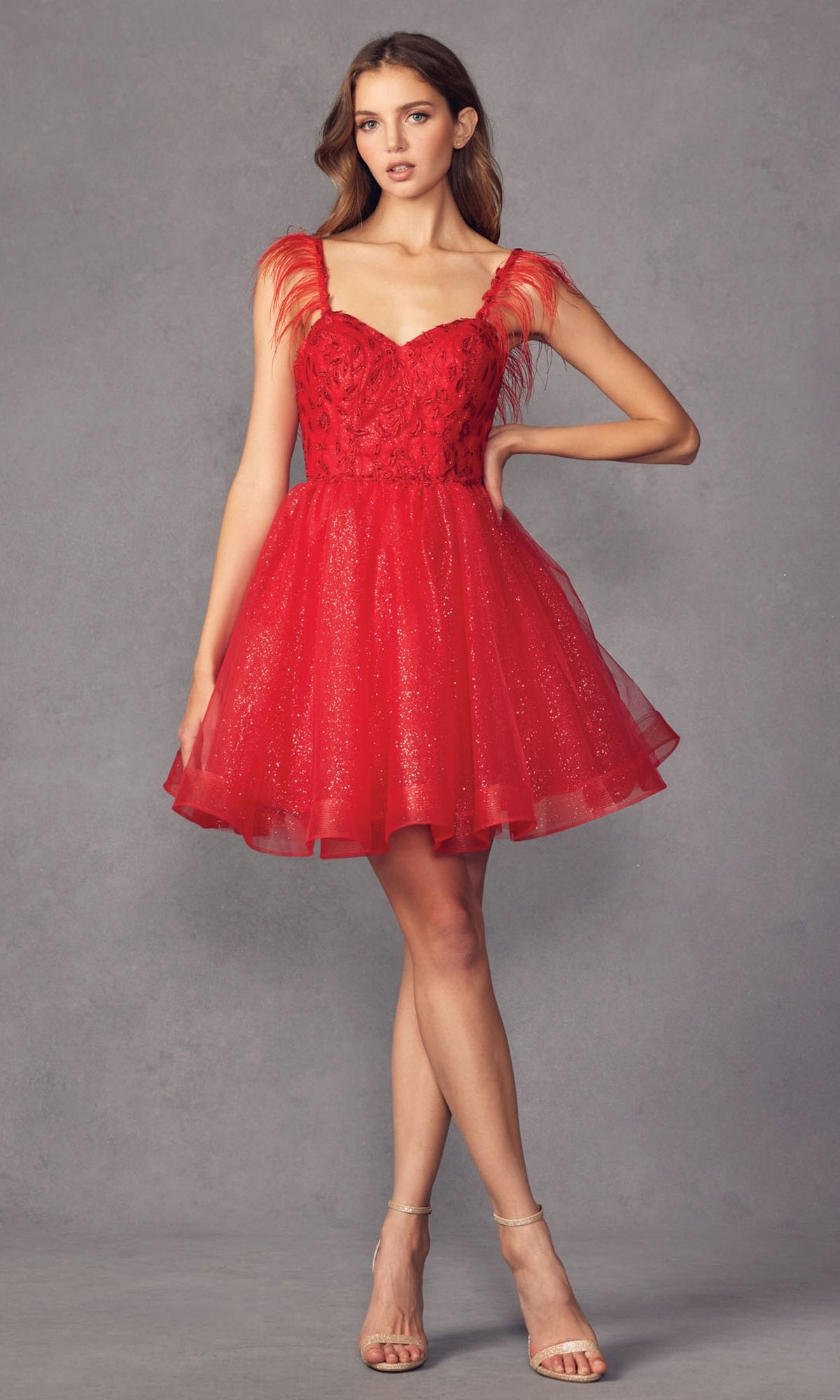 A woman wears the Short Babydoll Homecoming Dress with Feathers 881, featuring glitter fabric, feathered shoulder straps, and a flared skirt, as she stands against a plain gray background.