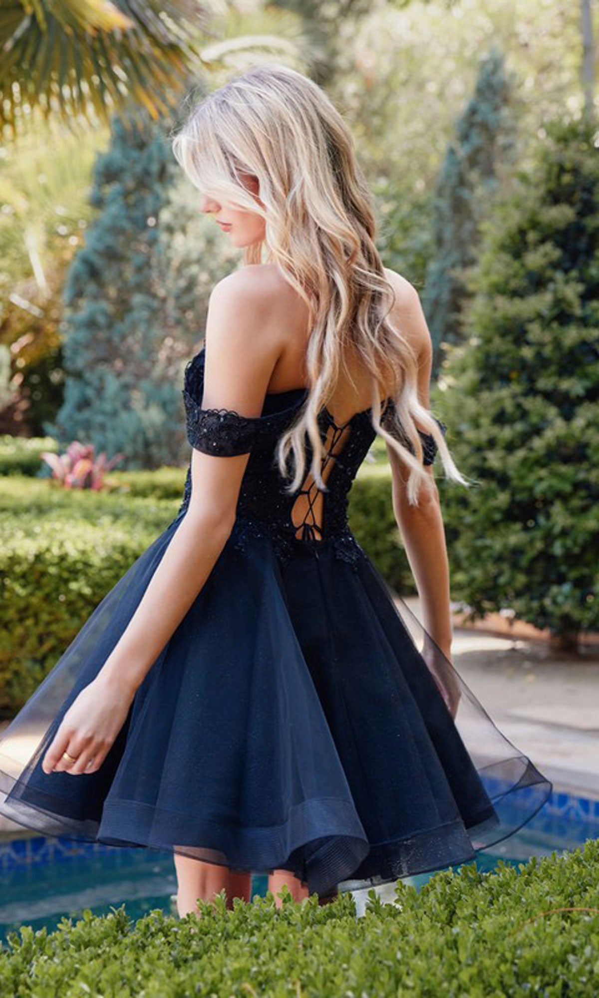 A woman with long blonde hair stands outdoors near manicured bushes and trees, wearing the Short Black Babydoll Homecoming Dress 880 featuring a lace-up back.