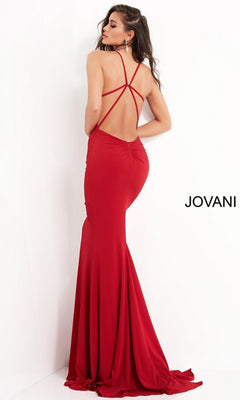 A woman models the Jovani Strappy-Back Long Jersey Prom Dress 00512, a floor-length red gown with crisscross straps and an open back, posing by a white wall featuring the Jovani brand name.