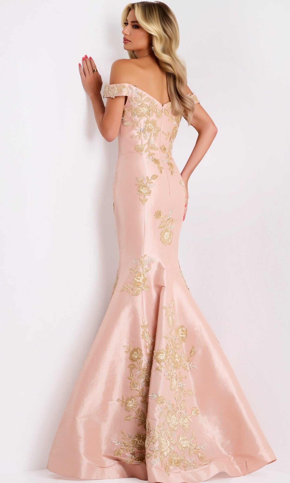 A woman in the Jovani Gold and Black Long Prom Dress 00635 stands facing away with her hand on her hip against a plain light background, striking a graceful pose that showcases the gown's elegant off-shoulder mermaid silhouette and gold detailing—perfect for a formal evening or prom.