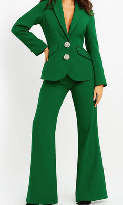 A person wearing the emerald Long Jumpsuit: Jovani 02637, which features flared pants and a blazer-style top with two decorative buttons, stands against a plain white background.