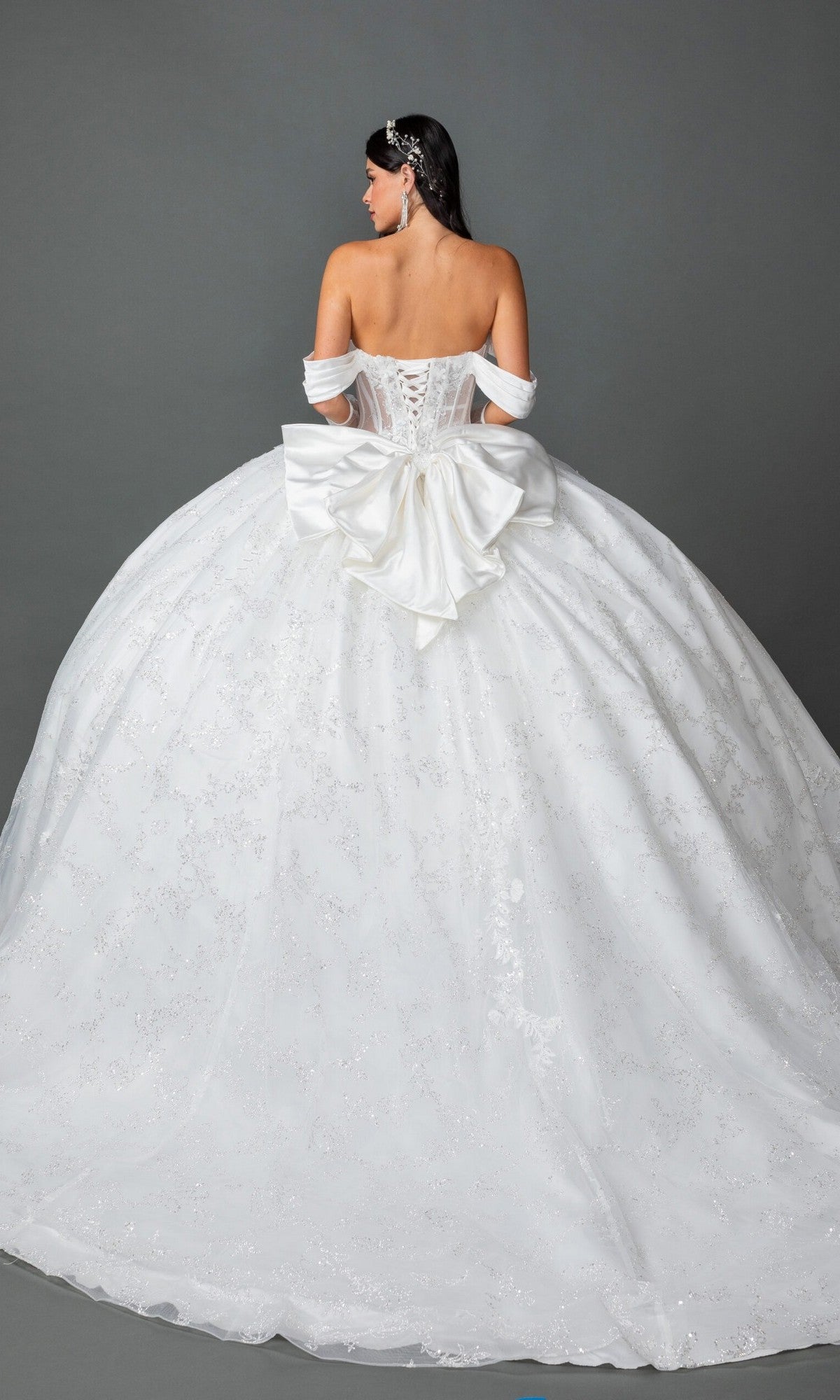 A woman faces away from the camera, wearing the Long Wedding Dress 0322 by Dancing Queen—an off-the-shoulder ballgown with a lace-up back, a large satin bow, and a full, embellished skirt—set against a plain gray background.
