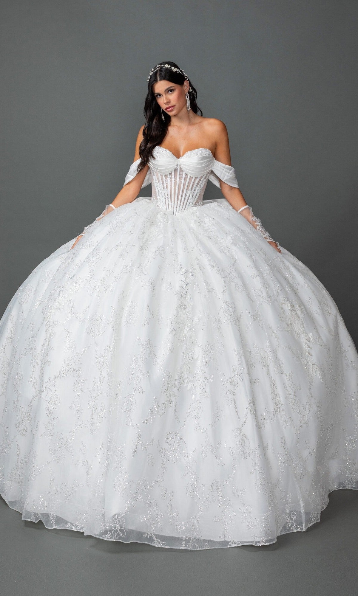A woman stands against a plain gray background wearing the Long Wedding Dress 0322 by Dancing Queen in off white, featuring an off-the-shoulder neckline, full skirt, illusion corset bodice, and a tiara.
