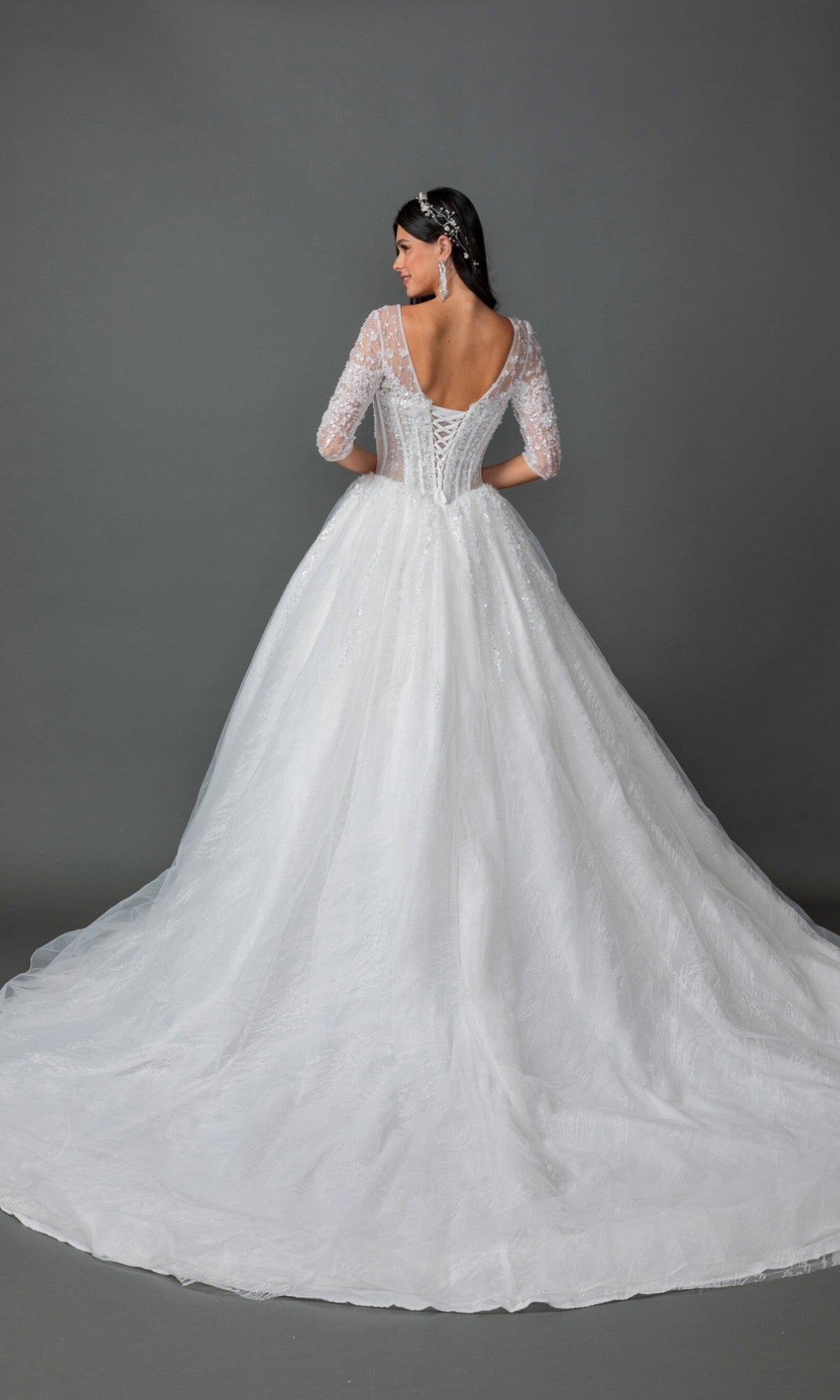 A woman stands facing away against a plain gray background, showcasing the intricate back detail of the Long Wedding Dress 0323 by Dancing Queen, which features a lace-embellished bodice and a full tulle ball gown skirt.