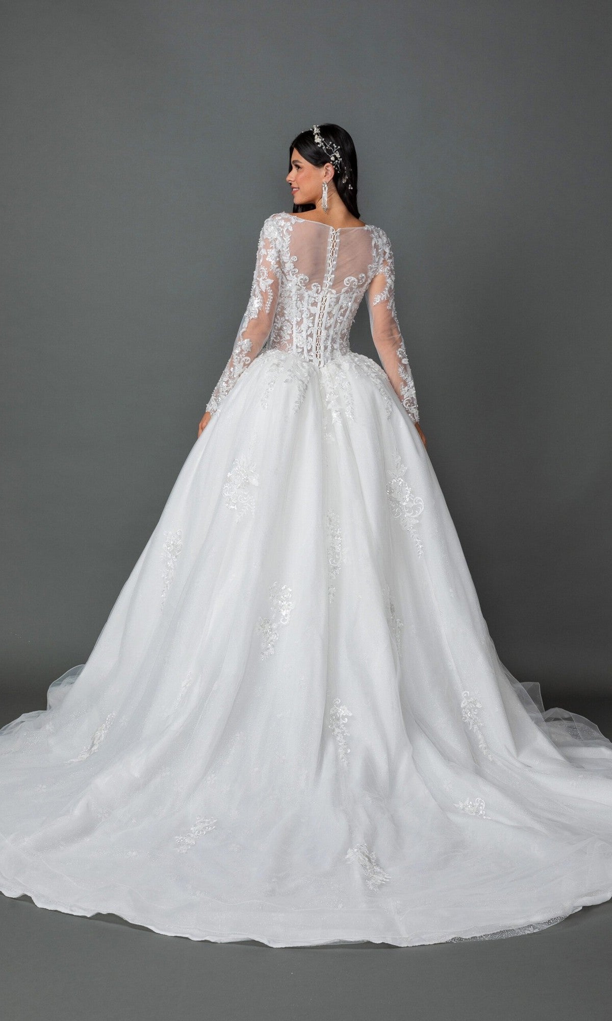 A woman faces away, wearing the Long Wedding Dress 0325 by Dancing Queen—a long-sleeved white gown embellished with delicate floral lace appliques, featuring a full A-line skirt and train—set against a plain gray background.