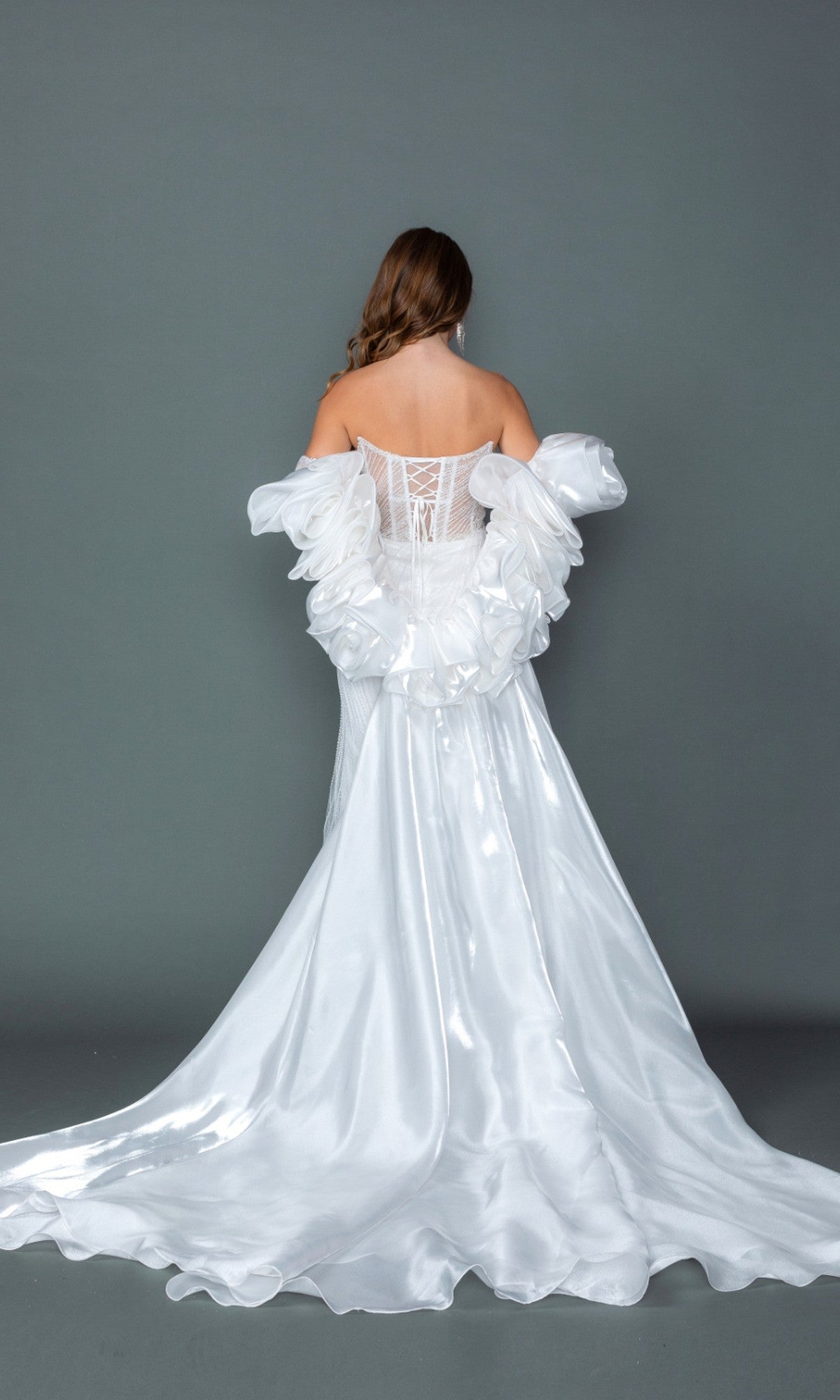 A woman stands facing away, wearing the Long Wedding Dress: Dancing Queen 0328, featuring a stunning white design with a strapless corset bodice, ruffled off-shoulder sleeves, and a shiny train, set against a plain gray background.