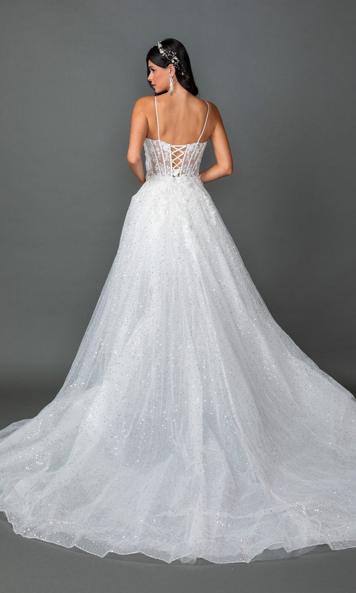 A woman stands facing away against a plain gray background, wearing the Long Wedding Dress 0329 by Dancing Queen—a sparkling white gown featuring a corset back, a long tulle train, and delicate floral lace appliques along the skirt that add an elegant touch.