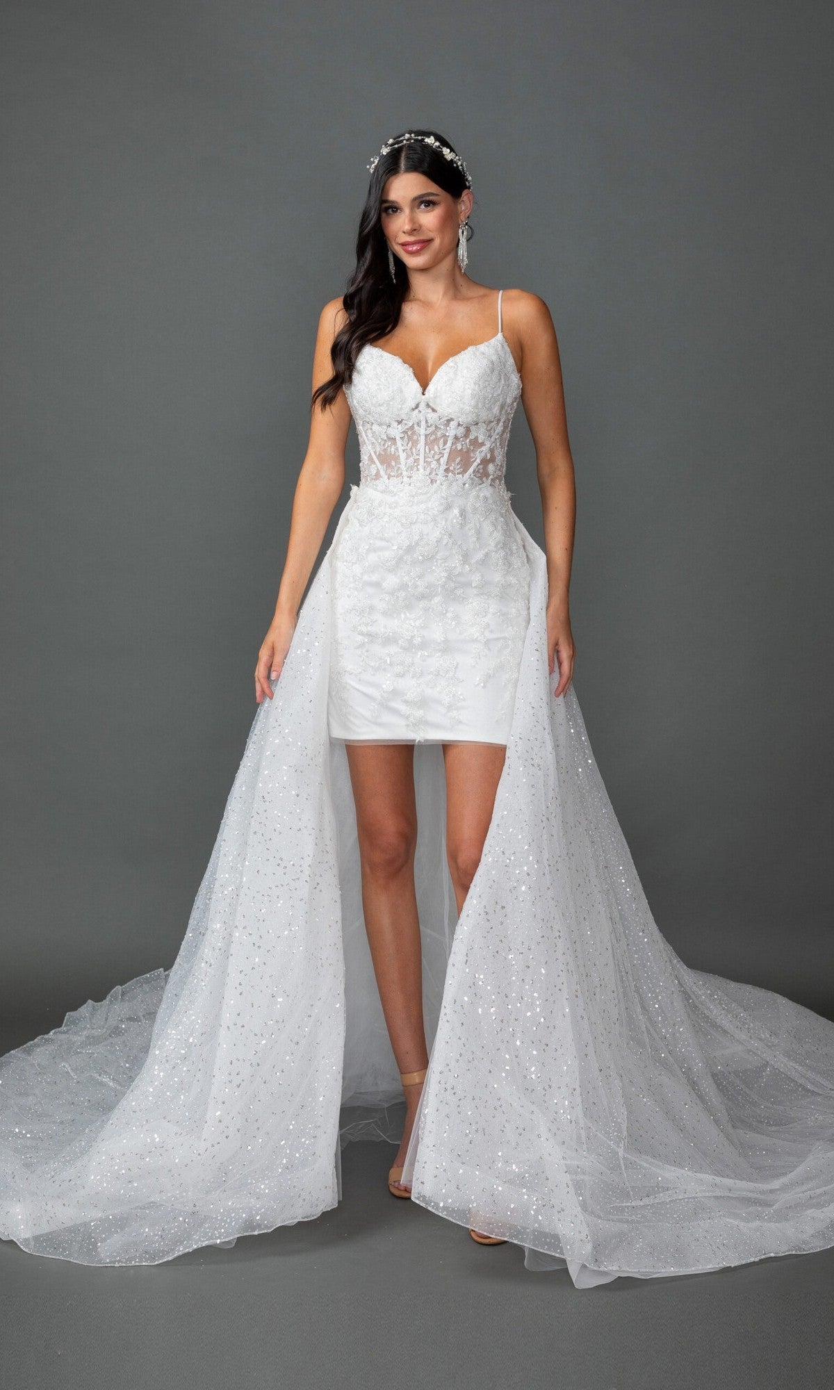 The woman is wearing the Long Wedding Dress 0329 by Dancing Queen in off white, featuring a sheer bodice with spaghetti straps, a short inner skirt, and an outer tulle train embellished with delicate floral lace appliques. She stands against a plain gray background.