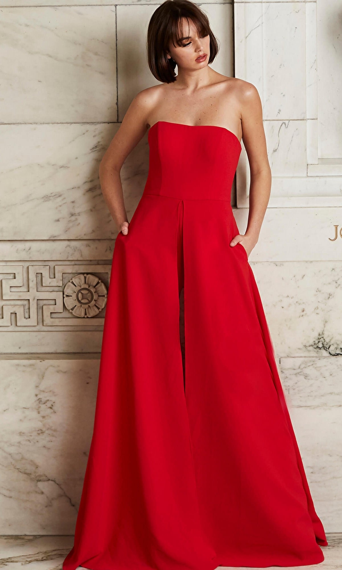 Woman wearing the Jovani Strapless Wide-Leg Formal Jumpsuit 03529 in red, featuring a strapless neckline, floor-length wide legs with a front slit, and pockets, stands against a marble wall.