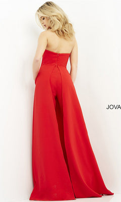 A woman stands facing a white wall, wearing the Jovani Strapless Wide-Leg Formal Jumpsuit 03529 in red, featuring a strapless neckline and floor-length wide legs.