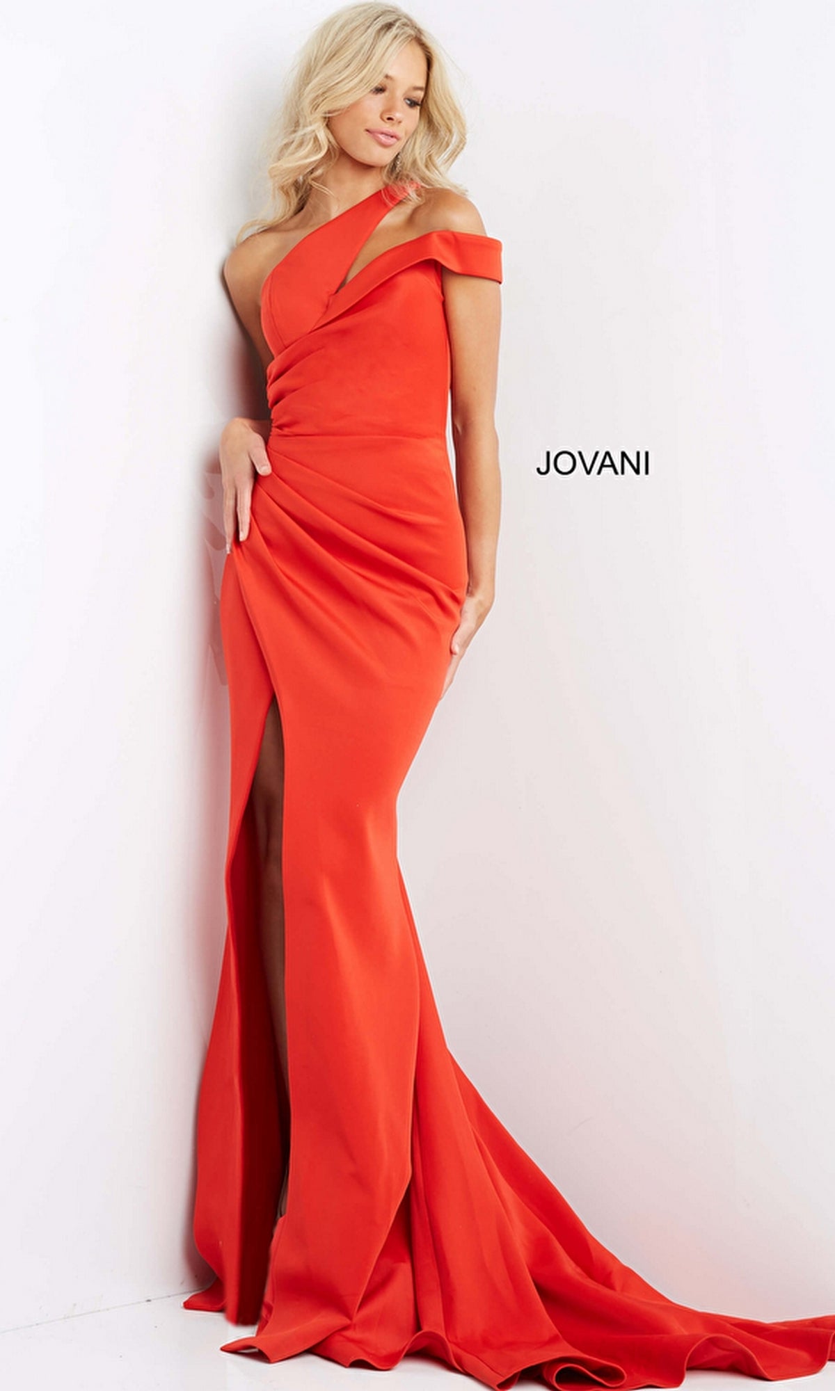 A woman wearing the Jovani 04222 One-Shoulder Long Mermaid Prom Dress in bright orange, featuring a thigh-high slit, stands against a white wall with 