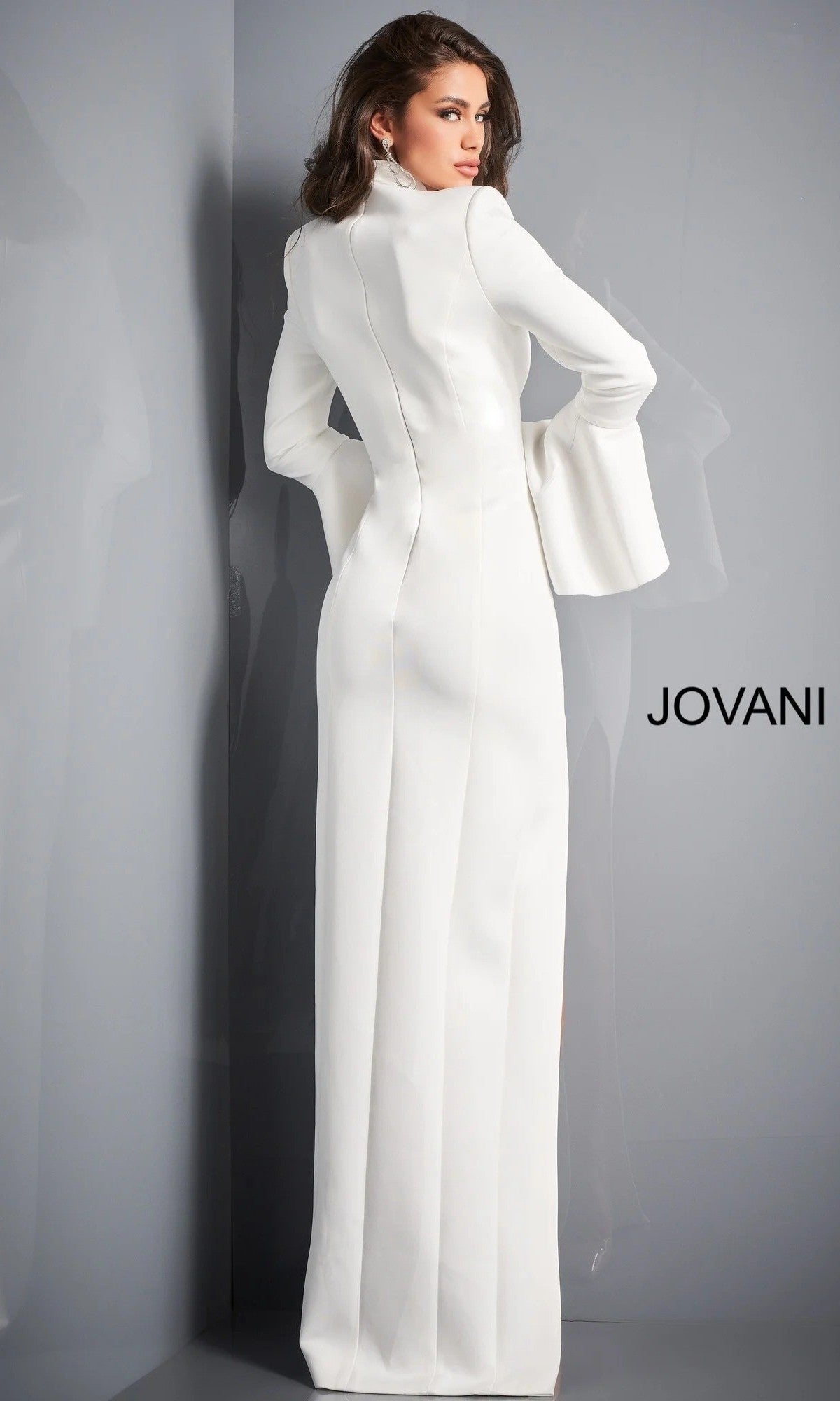 A woman stands facing away, her head turned to the side, wearing the Long Formal Dress: Jovani 04240—a white, long-sleeved, floor-length gown—next to a gray wall featuring the “JOVANI” logo.