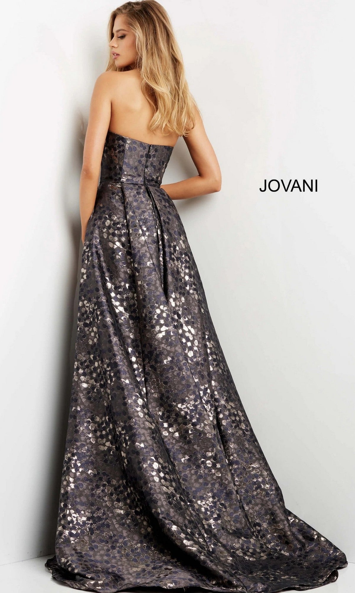 A woman stands facing a wall beside the inscription “JOVANI,” wearing the Jovani 06255 long formal dress—a strapless, floor-length metallic jacquard gown featuring a floral pattern.