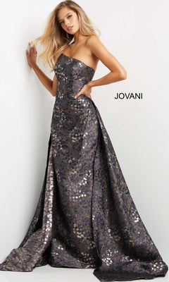 A woman poses against a white wall wearing the Long Formal Dress: Jovani 06255, a strapless jacquard gown featuring a floor-length metallic-patterned design with a subtle flare and lustrous shine. The word “JOVANI” is displayed beside her, capturing the refined elegance of this designer creation.