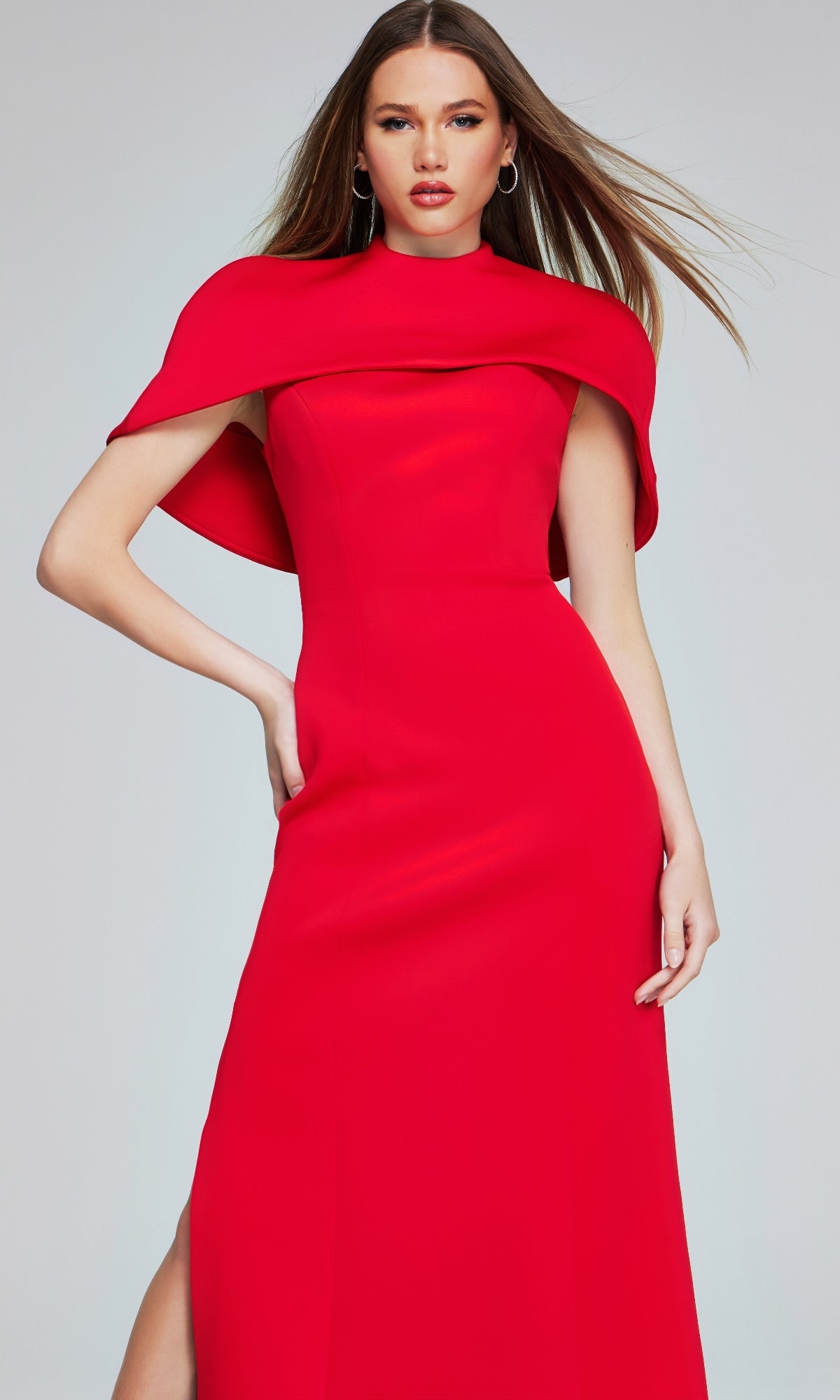 A woman wearing the Jovani 06538 long formal dress, a sleek floor-length red gown with elegant cape detail, stands against a plain light background.