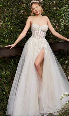 A woman stands outdoors against lush greenery, wearing the Long Prom Dress 06610 by Jovani—an off white strapless corset gown featuring sheer floral lace details and a thigh-high slit.