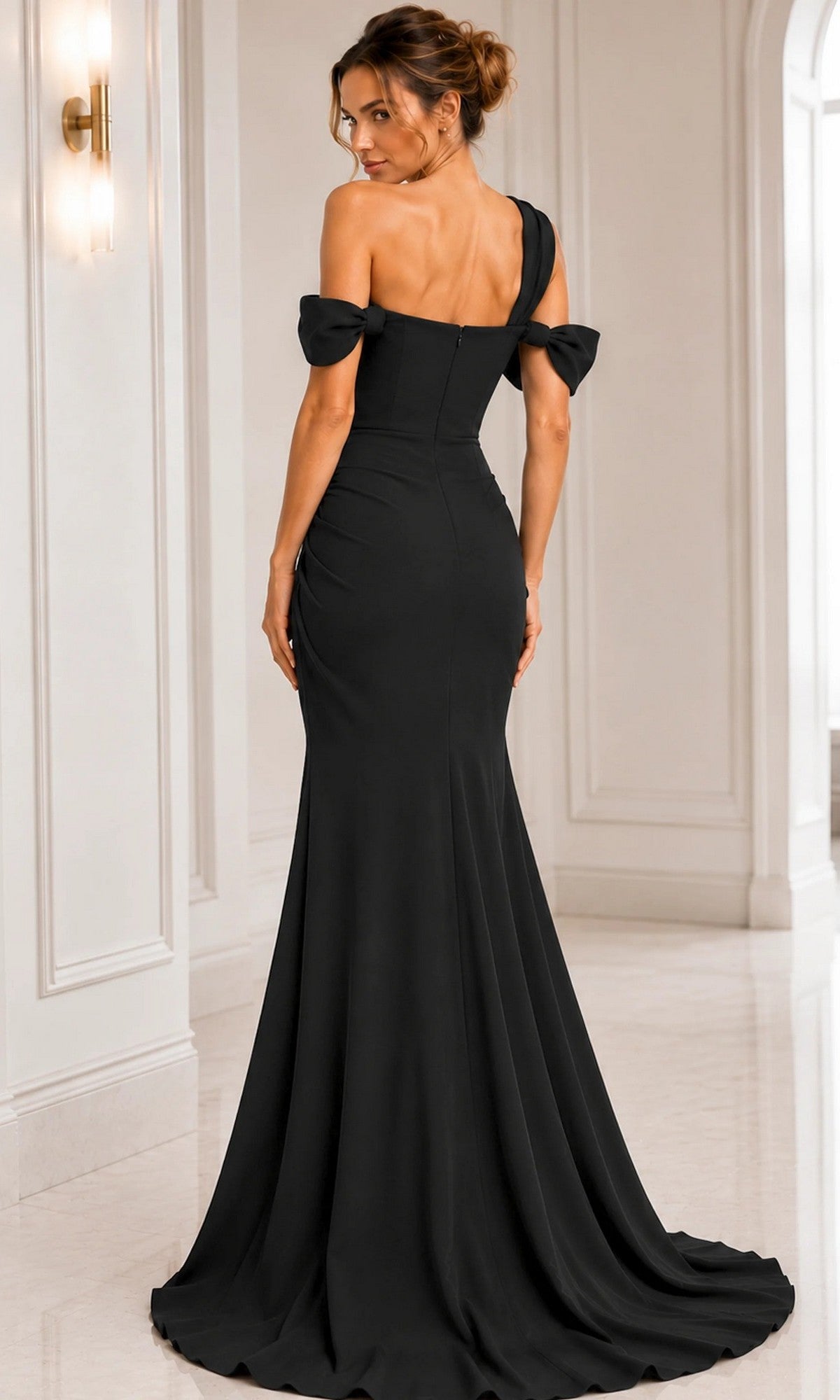 A woman stands facing away from the camera in a white, elegant room with light marble flooring, wearing the Jovani 06723 long formal dress—an off-shoulder, floor-length black gown.