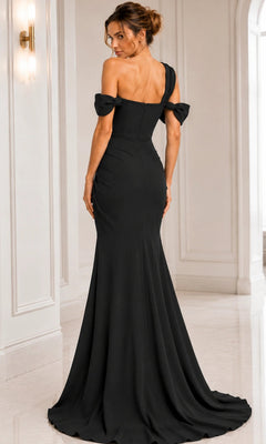 A woman stands facing away from the camera in a white, elegant room with light marble flooring, wearing the Jovani 06723 long formal dress—an off-shoulder, floor-length black gown.
