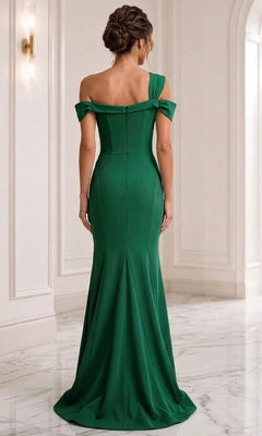A woman stands in an elegant white room with marble floors, facing away from the camera, wearing the Long Formal Dress: Jovani 06723—a fitted, floor-length green gown featuring off-the-shoulder straps and a graceful sheath silhouette.