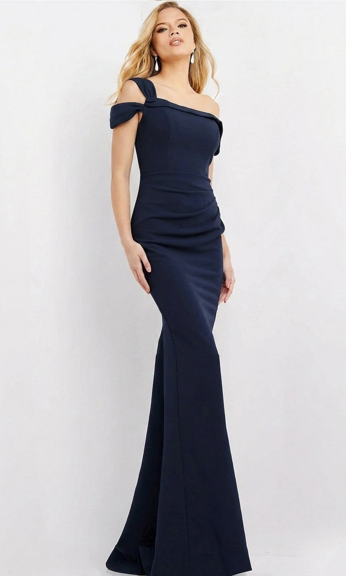 A woman stands against a plain light background wearing the Jovani 06723 long formal dress in navy, which features a fitted, off-the-shoulder design and a sleek sheath silhouette, capturing its timeless elegance.