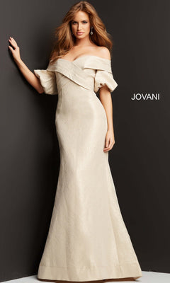 A woman stands against a dark wall wearing the Jovani 06831 long formal dress in champagne—a floor-length, off-the-shoulder gown with a fitted mermaid silhouette and textured fabric. The word “JOVANI” appears on the wall behind her.