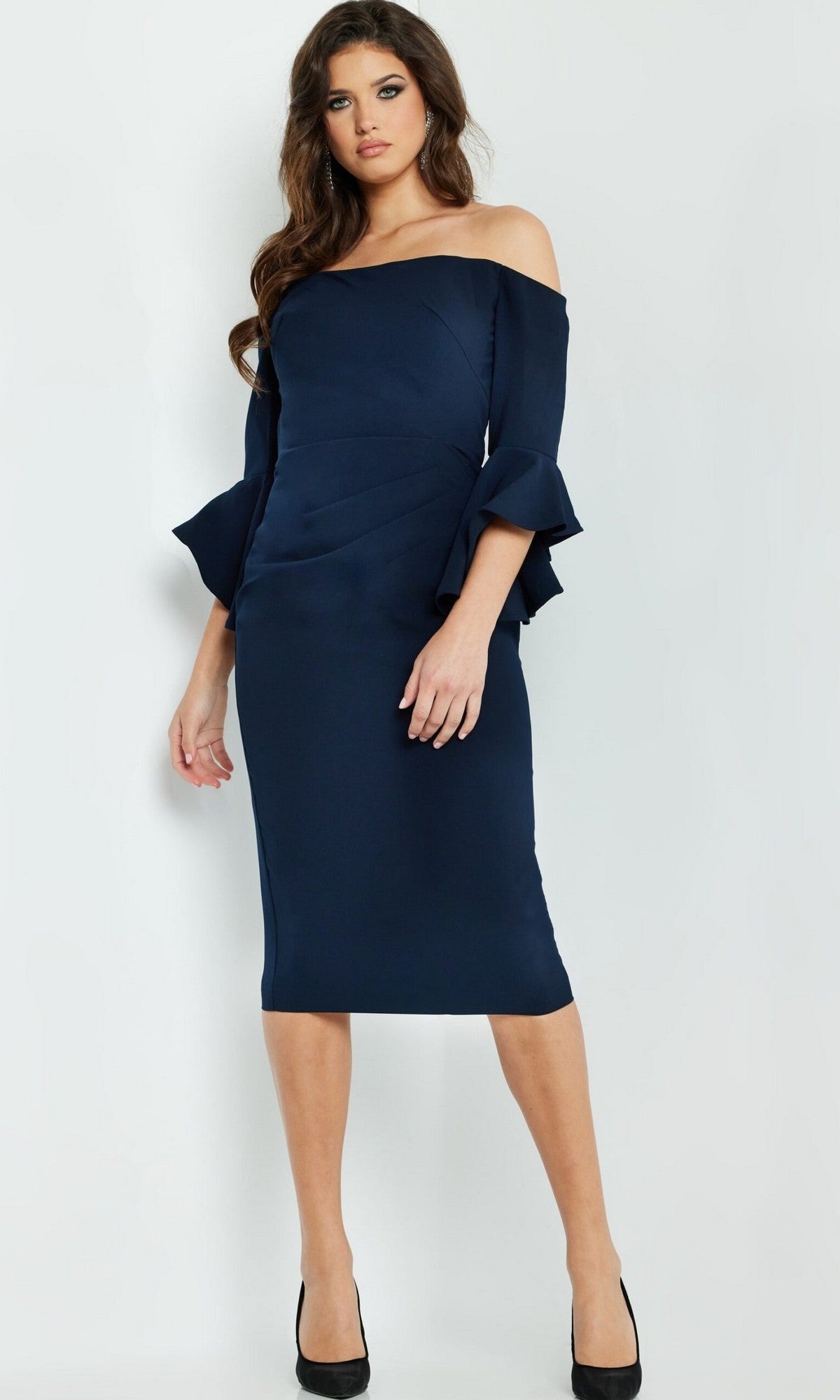 A woman stands against a plain background wearing the Jovani 07066, a navy blue off-the-shoulder crepe sheath dress featuring knee-length styling and flared sleeves—an elegant choice as a short wedding guest dress—paired with black high heels.