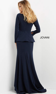 A woman stands with her back to the camera, modeling the Jovani 07131 long formal dress, which features elegant long sleeves, a floor-length trumpet silhouette, and sophisticated peplum detailing. The brand name “Jovani” is visible on the wall behind her.