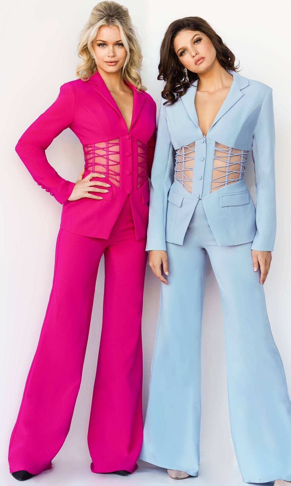 Two women stand side by side wearing the Jovani 07227 long jumpsuit, featuring tailored silhouettes with lace-up corset details and a touch of sheer at the waist; one is in vibrant pink, while the other is in soft light blue.