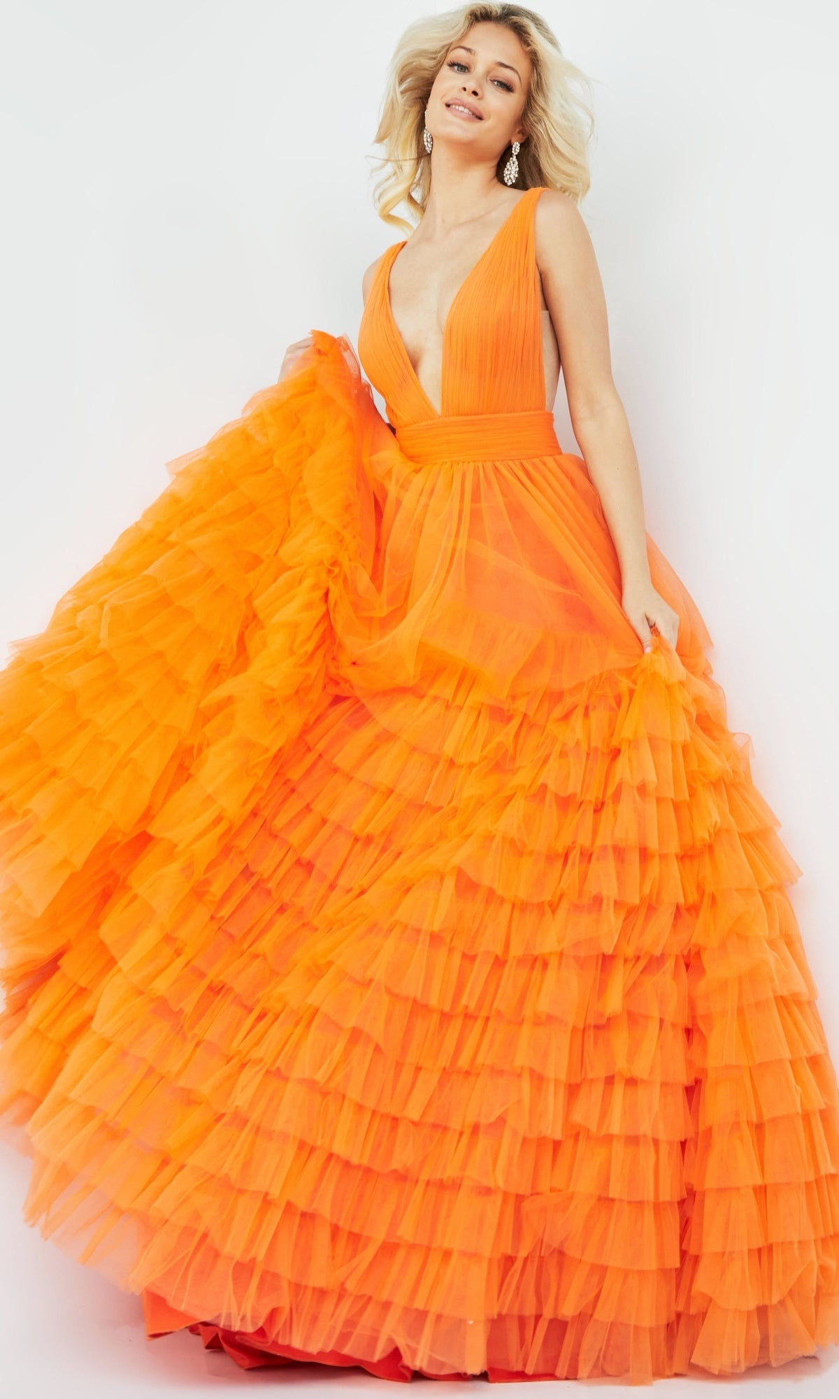 A woman smiles and poses in the Jovani V-Neck Long Ruffled Prom Ball Gown 07264, featuring bright orange fabric, dramatic ruffles, and a deep V-neckline against a plain background.