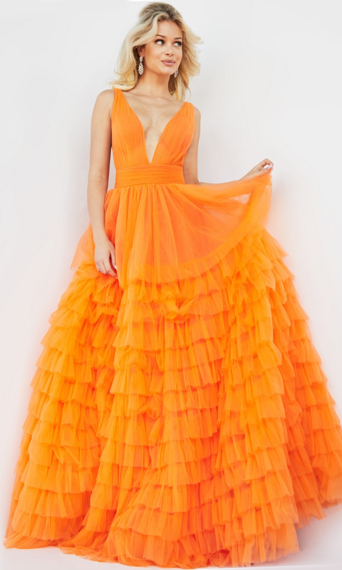 A woman wears the Jovani V-Neck Long Ruffled Prom Ball Gown 07264, a sleeveless bright orange dress with a deep V-neck and a full ruffled tulle skirt, standing against a plain light background.