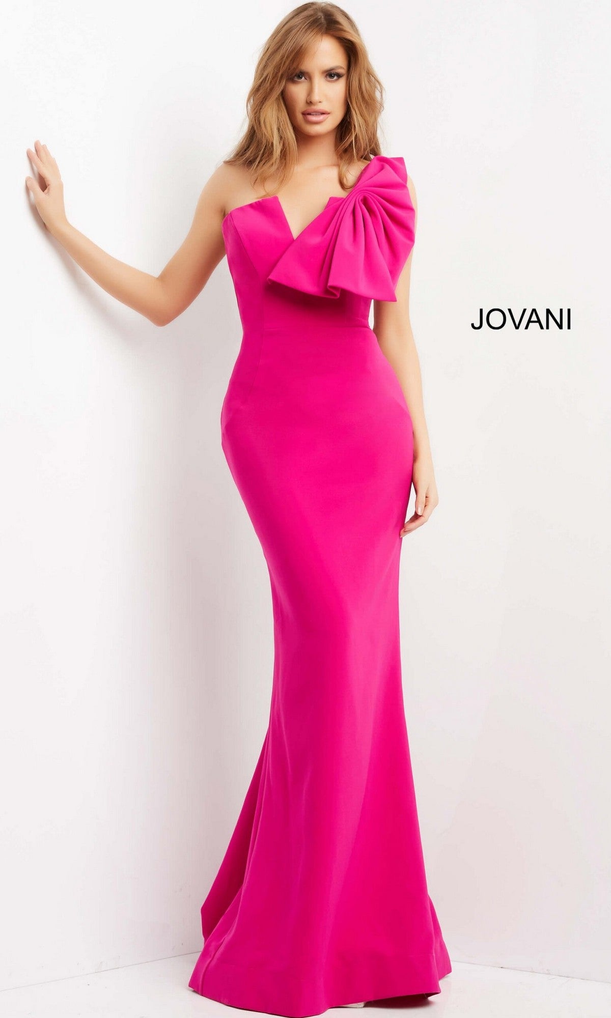 A woman stands against a white wall wearing the Jovani 07306 long formal dress in fuchsia, featuring a floor-length one-shoulder design with a large bow detail on the shoulder.